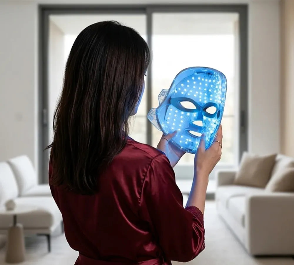 Mulher em pé utilizando a máscara de LED azul próxima à janela demonstrando leveza do aparelho Loja Morela