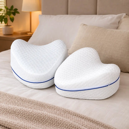 Dois travesseiros anatômicos brancos em formato de coração para joelhos e pernas apoiados na cama, destacando o formato ergonômico e o zíper azul.
