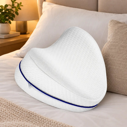 Travesseiro ortopédico anatômico branco com zíper azul posicionado sobre uma cama arrumada com lençóis brancos e iluminação aconchegante.